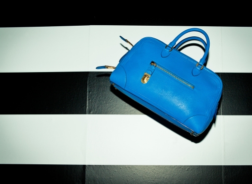 Bright blue leather handbag on a black and white striped tile floor with a gold-tone zipper and lock for a fashion editorial shot.