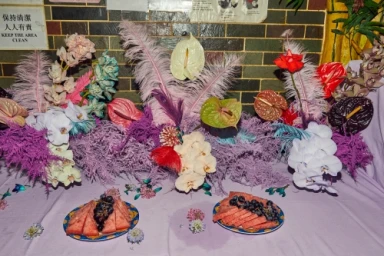 An elaborate floral arrangement with orchids, anthuriums, and feathers above two plates of watermelon and grapes on a lavender tablecloth.