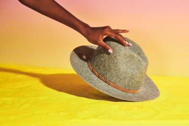 Close-up of a dark-skinned hand resting on the brim of a grey felt fedora hat with a brown leather band on a yellow surface.