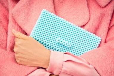 Close-up of a person holding a bright blue beaded clutch handbag against a textured pink faux fur coat and pink shirt.
