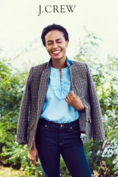 Smiling woman wearing a tweed blazer, light blue ruffled blouse, and dark denim jeans outdoors.
