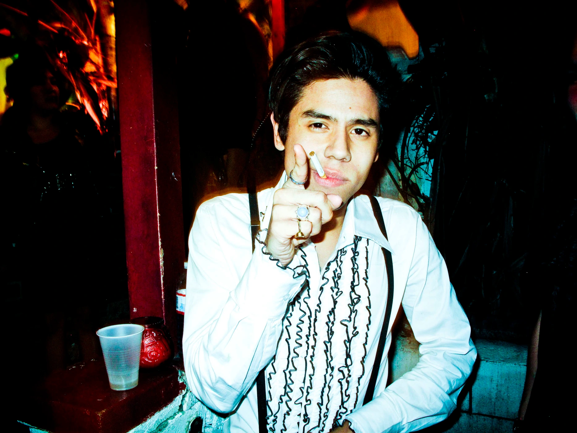 Young man in a white ruffled shirt and suspenders pointing forward with a cigarette in his mouth by photographer Meredith Jenks.
