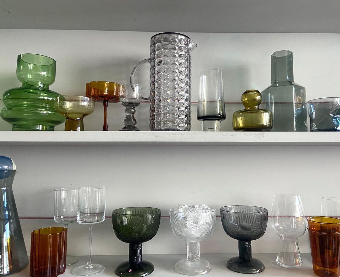 Assorted collection of colored glass vases and drinking glasses displayed on two white shelves against a plain, light wall.