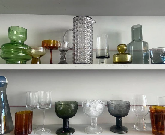 Assorted collection of colored glass vases and drinking glasses displayed on two white shelves against a plain, light wall.