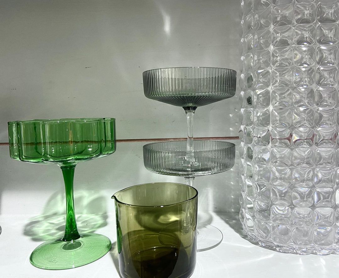 Green and clear glassware on a reflective white surface in a still life arrangement, including a pedestal dish and a textured vase.