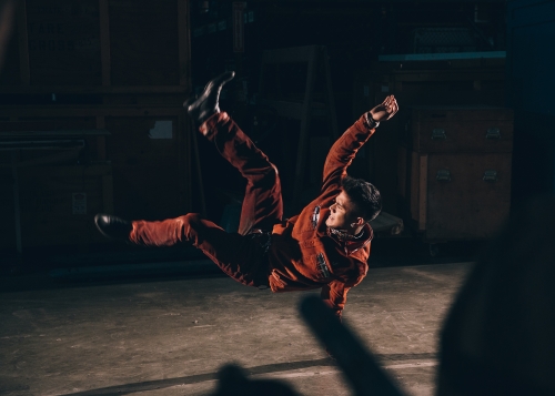 Man in red tracksuit performing a dance move, mid-air, in a dimly lit industrial or studio setting. Dynamic action shot.