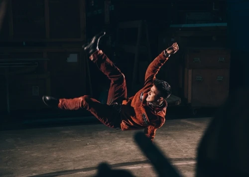 Man in red tracksuit performing a dance move, mid-air, in a dimly lit industrial or studio setting. Dynamic action shot.