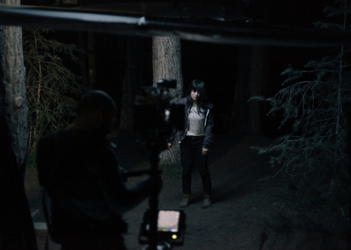 Woman with dark hair stands in a forest, possibly during a film shoot, with a camera in the foreground and trees in the background.