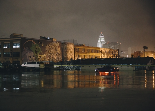 Cityscape at night with water reflecting buildings, illuminated by streetlights and car headlights creating a moody urban scene.