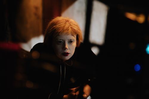 Portrait of a fair-skinned adult female with orange hair, red lipstick, and white eyelashes posing indoors in front of dark background.