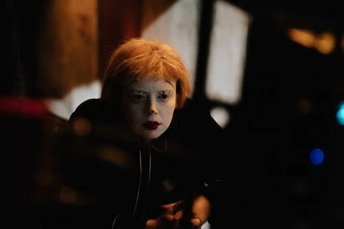 Portrait of a fair-skinned adult female with orange hair, red lipstick, and white eyelashes posing indoors in front of dark background.