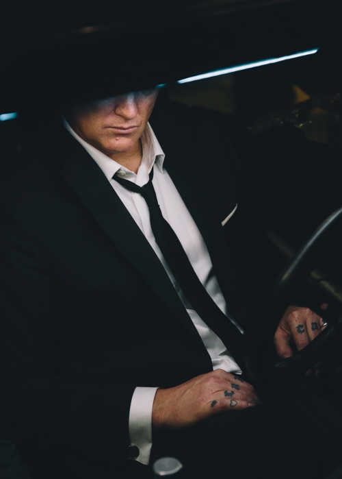Stylish man in suit sits in car wearing dark hat. The individual has hand tattoos, white dress shirt, and tie in low-key lighting.