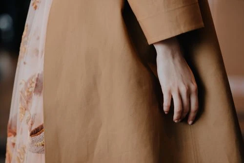 Close-up of a woman's hand and forearm resting against a brown trench coat, with a floral dress peeking out from underneath.