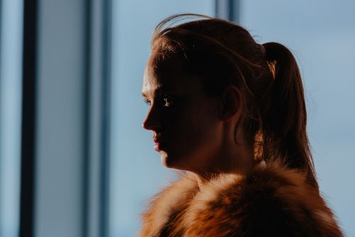 A blonde woman in a black top poses in front of a window with sheer white curtains, bathed in soft, natural light, for a portrait.