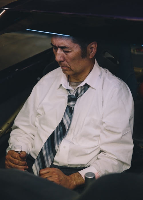Middle-aged man in white shirt and patterned tie sitting inside a car holding a bottle, photographed by Joyce Kim.