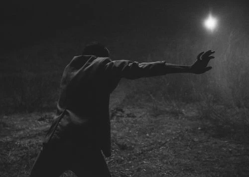 Monochromatic image featuring an adult male reaching toward a bright light outdoors in a dark, rural setting, evoking mystery.