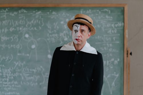 Adult male in a dark coat and hat with clown makeup stands against a green chalkboard covered in chalk math equations indoors.