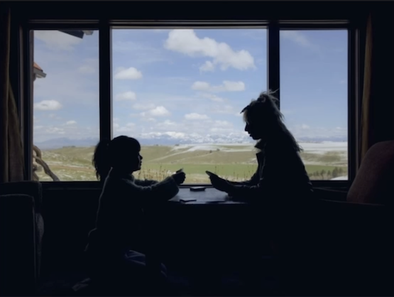 Silhouette of an adult and a child sitting at a table indoors, looking out a window at a rural landscape with distant mountains.