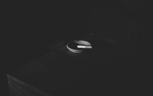 Monochromatic image of a lit cigarette resting in an ashtray, centered on a dark wood surface, creating a somber, minimalist composition.
