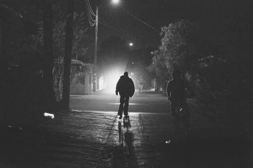 Monochrome, low-angle night shot of two people walking on a dark street, illuminated by streetlights creating long shadows.