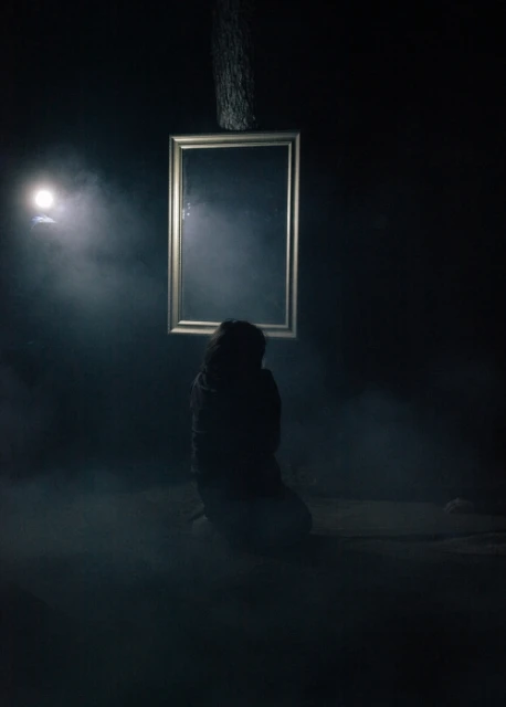 Person kneeling in a dark, smoky room with a framed mirror and a single spotlight shining from the side, creating a moody scene.
