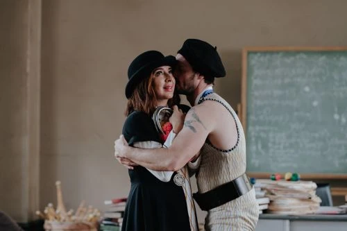 Couple in vintage attire embraces in a room with books and a chalkboard, the man kissing the woman's cheek in soft, diffused lighting.