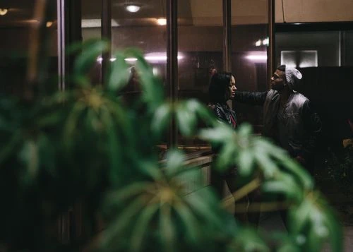 Couple stands outside glass building with green plant foreground. Adult male wears knit hat, adult female has shoulder-length hair.