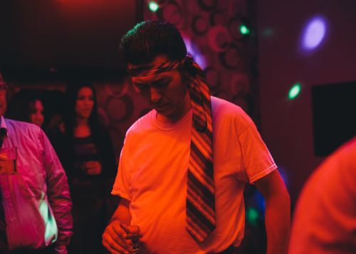 Man in white t-shirt and tie around his head, holding a glass in a dimly lit room with other people in the background.