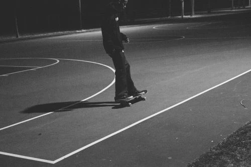 A skateboarder practices at night on an outdoor court with white lines, illuminated by dim light, creating a moody, monochromatic scene.