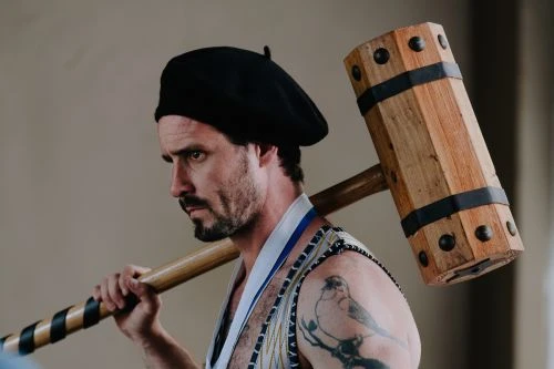 Adult male with a beard and black hat carries a large wooden hammer. He is looking to the side in an indoor setting.