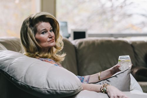 Mature woman relaxes on a couch with a drink. She has blonde hair and is wearing a patterned top and gold bracelets, looking thoughtful.