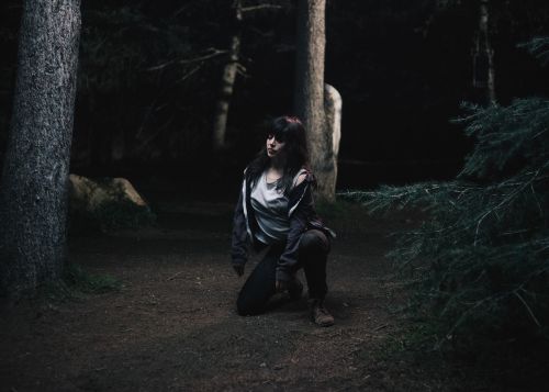 Woman kneeling in dark forest, wearing a grey jacket and dark pants, surrounded by trees and foliage in a moody, natural environment.