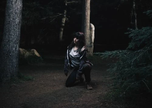 Woman kneeling in dark forest, wearing a grey jacket and dark pants, surrounded by trees and foliage in a moody, natural environment.