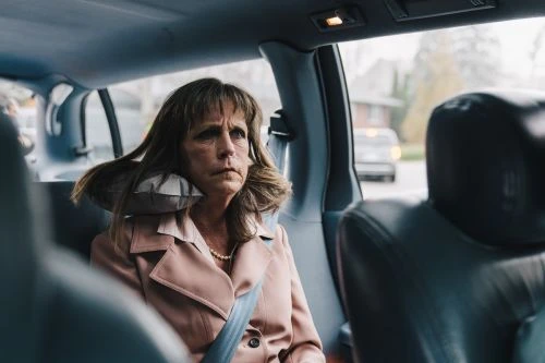 Mature woman in a pink jacket and seatbelt rides in a car with a pillow behind her head, looking thoughtfully out of the window.