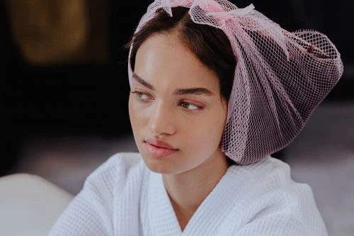 Close-up of a young woman with light skin wearing a white robe and a pink head covering, looking to the side in an indoor environment.