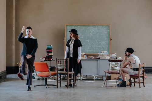 Three people in an indoor setting with a vintage aesthetic, featuring a chalkboard, books, and mismatched furniture creating a staged look.