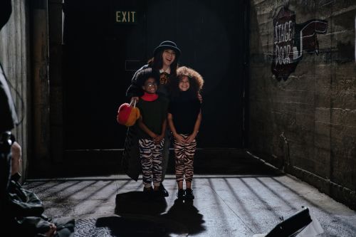 A woman and two children stand in a dimly lit alleyway with concrete walls and metal bars, posing for a photograph in a backstage area.