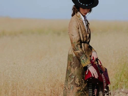 A woman wearing an embroidered tan coat and black hat stands in a grassy field holding a dark red handbag in an outdoor setting.