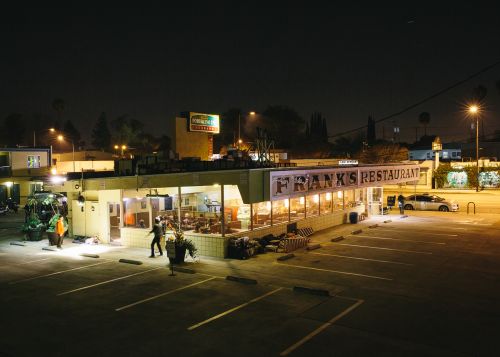 Night scene of Frank's Restaurant, illuminated with warm light, shows a classic diner setup with people, parked cars, and streetlights.