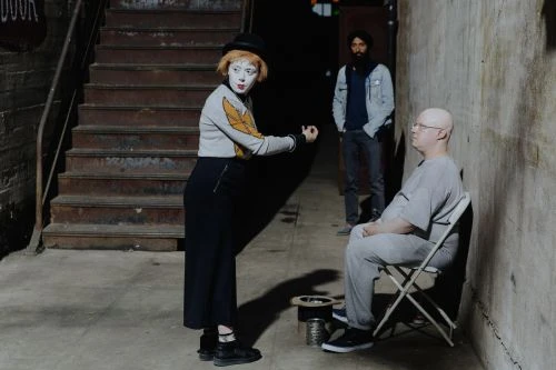 Mime artist in black hat, exaggerated white makeup, grey patterned sweater gesturing to seated adult male with a bearded man behind them.