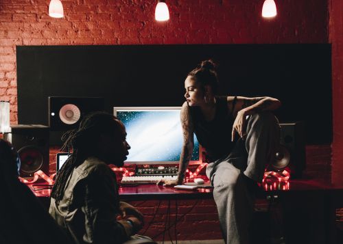 In a music studio, a woman sits on a desk near a computer monitor, while another person sits in a chair in a dimly lit recording space.