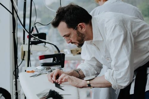 Bearded man in white shirt working at a tailoring table with scissors and an iron by Joyce Kim