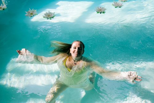 Woman with long hair floats in turquoise water with white water lilies, wearing a white dress, and smiling.