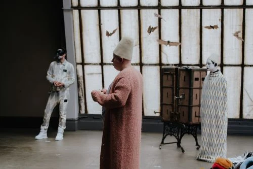 Three people stand in a room with tall windows as paper bats hang in the foreground. One wears white makeup and a patterned garment.