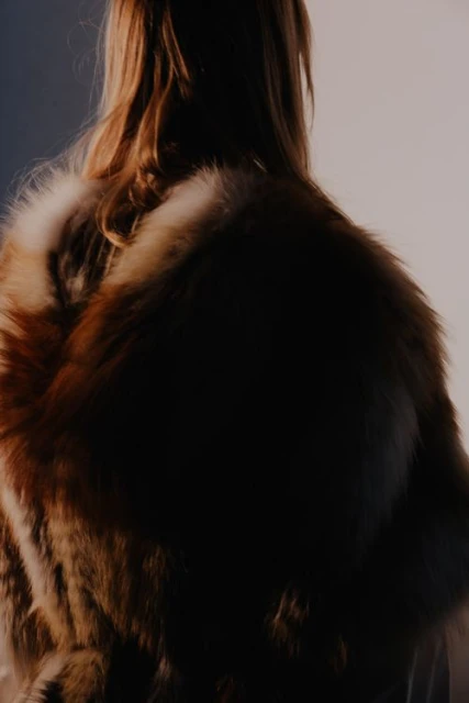 Back view of a woman with auburn hair wearing a fur coat in front of a split gray and white backdrop with natural lighting.