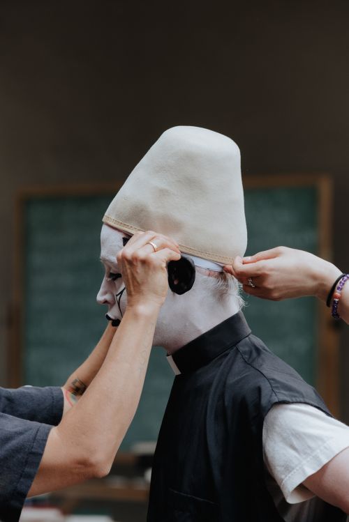 Person with white face paint and pointed hat having hat adjusted, with blurred background, art and fashion-forward, indoors.