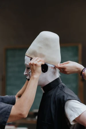 Person with white face paint and pointed hat having hat adjusted, with blurred background, art and fashion-forward, indoors.