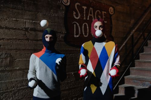 Two performers in colorful sweaters, black face coverings, juggling white snowballs on an indoor stage with a vintage stage door sign.