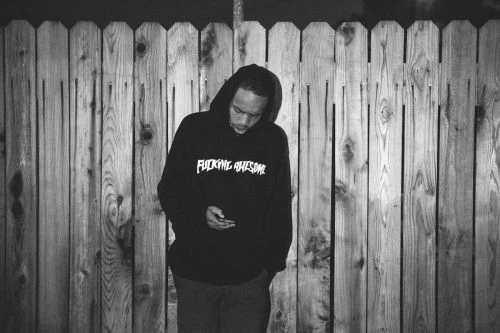 Monochrome photo of a person in a hoodie looking at a phone in front of a weathered wood fence, creating a textured, urban scene.