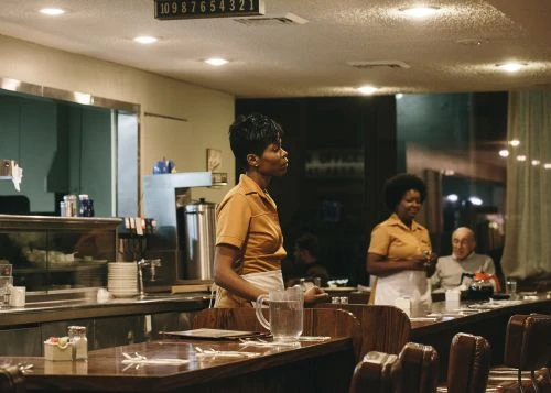 Two waitresses in yellow uniforms stand at a counter in a retro diner, serving a customer. Tables and chairs fill the foreground.